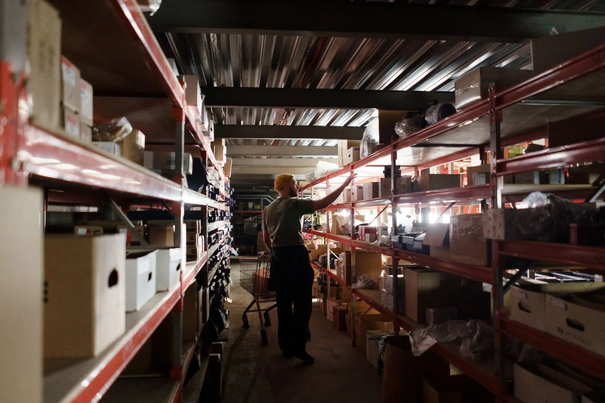 Worker reaching up to restock boxed parts on long metal shelving in a dim storeroom, the kind of stock a supplier may still own until it is paid for
